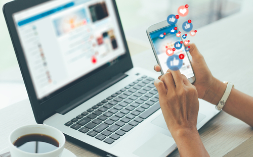 Picture of a female on an open laptop with the facebook app pulled up and the facebook app pulled up on her smartphone. There is a cup of coffee sitting next to the laptop on the table.