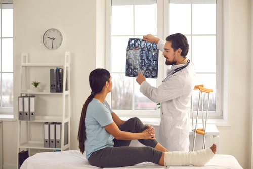 Picture of a female patient sitting on an examine table. She is wearing a leg cast. There is a male Physician standing next to her showing her X-rays.