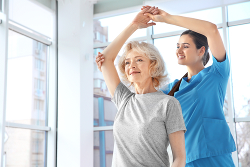 Picture of a female Rehabilitation Therapist standing behind an older female patient who has her arm stretched over her head. The Therapist is holding the patients elbow and her hand up over her head giving her support.