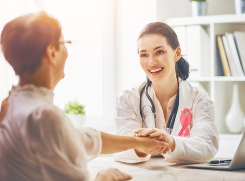 Picture of a female Physician sitting down at a desk with a elderly female patient. She is smiling at the patient with both of her hands holding the patients hand.