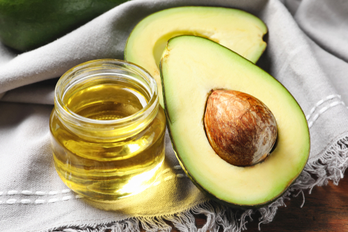 Picture of Avocado oil in a small glass container with a split Avocado sitting next to it on a cloth that is sitting on top of a table.