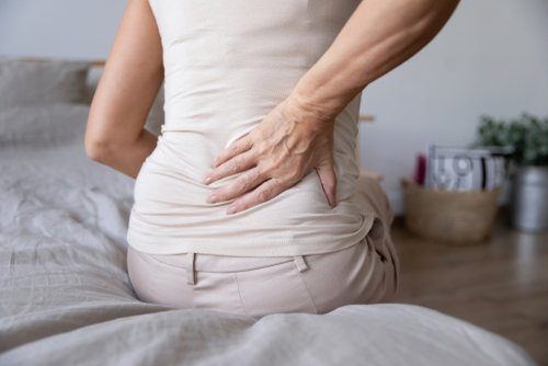 Picture of a female sitting on the edge of a bed holding her lower back with one hand.