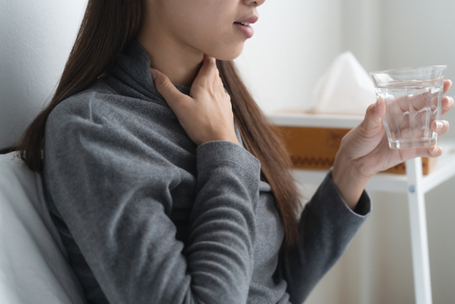 Picture of a woman holding her throat with one hand and holding a glass of water with the other hand.